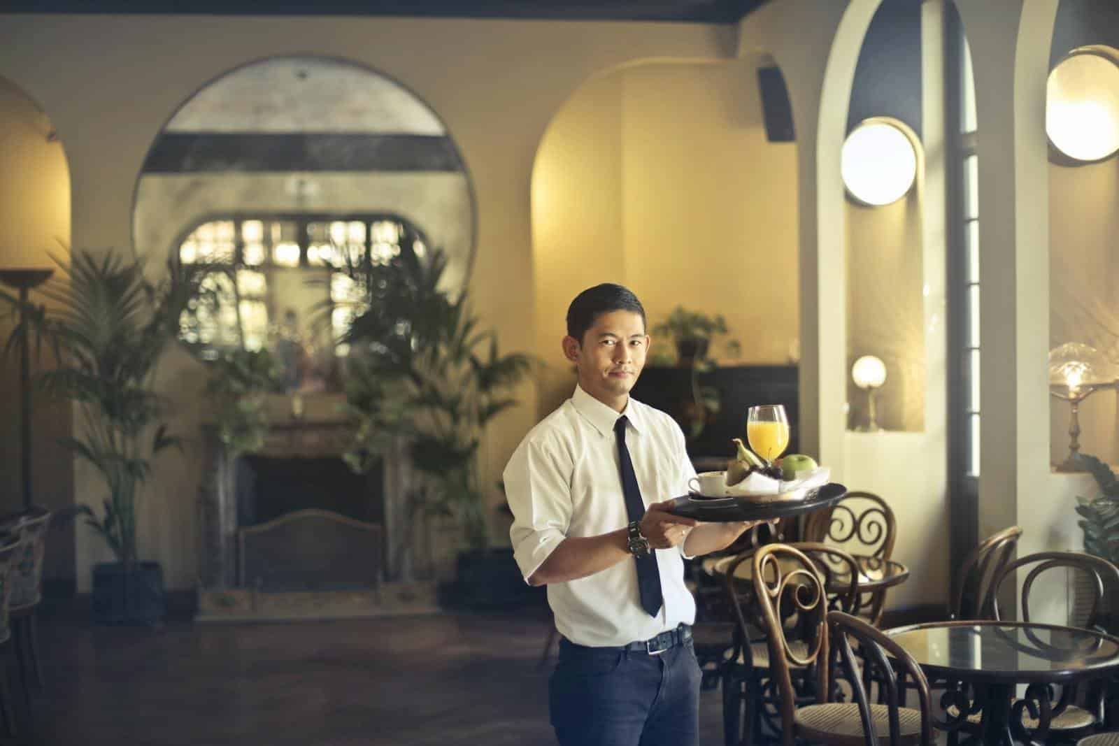 Confident young ethnic waiter in elegant clothes holding tray with food and drinks and looking at camera while serving tables in stylish restaurant