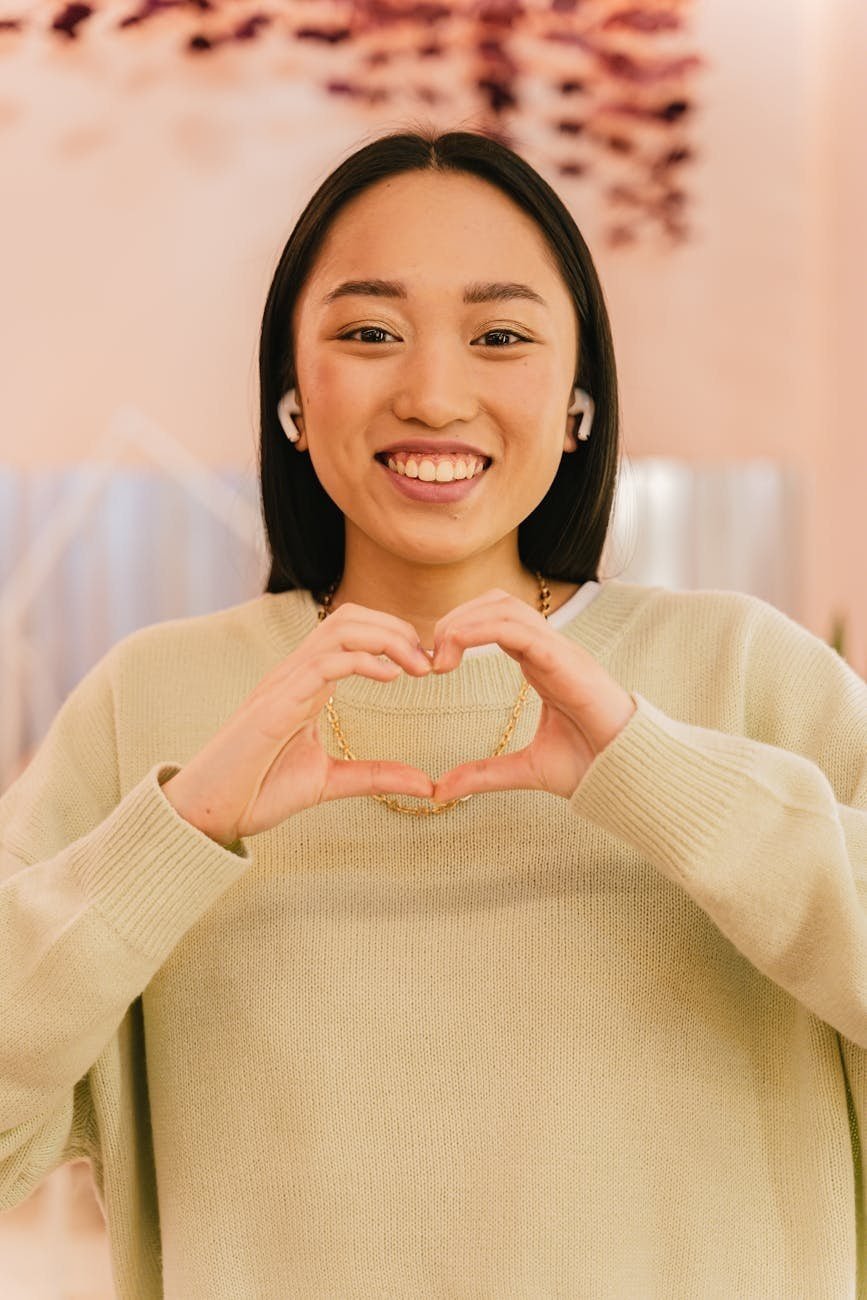 woman in beige sweater and wireless earphones smiling and showing heart shaped palms